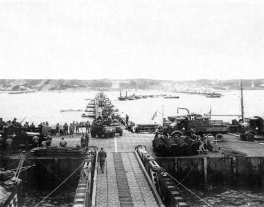 Veículos militares (como M3 Halftracks, um M8 Greyhound e AFKWX Trucks) aproximam-se da costa no porto artificial de Mulberry, através de uma ponte flutuante, para Omaha Beach.