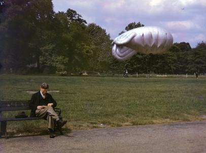 Um homem sentado num banco de jardim, em Londres, lendo um livro. Também um "balão barragem" é visível no fundo, enquanto um segundo voa alto a alguma distância.